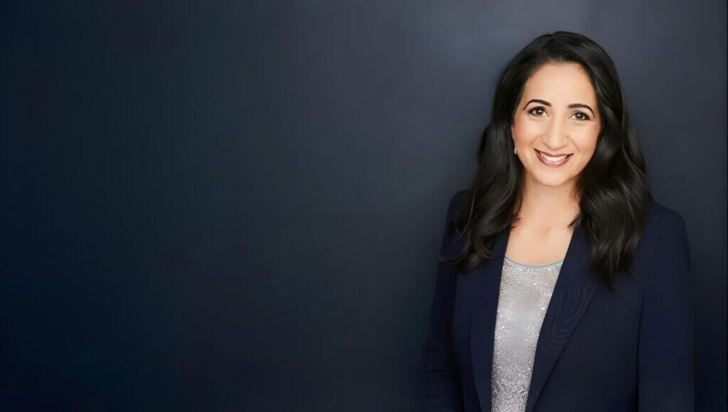 Headshot of a smiling woman with long dark hair in a navy blazer against a dark background